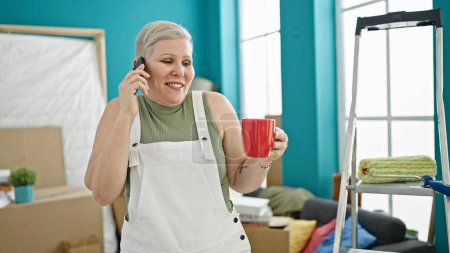 Foto de Mujer de pelo gris de mediana edad hablando en el teléfono inteligente bebiendo café en un nuevo hogar - Imagen libre de derechos