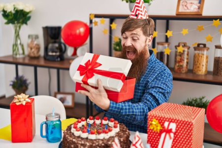 Téléchargez les photos : Jeune rousse homme célébrant anniversaire déballer cadeau à la maison - en image libre de droit