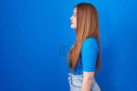 Téléchargez les photos : Femme rousse debout sur fond bleu regardant vers le côté, pose de profil relax avec le visage naturel et sourire confiant. - en image libre de droit