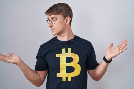 Téléchargez les photos : Homme blond caucasien portant Bitcoin t-shirt souriant montrant les deux mains paumes ouvertes, présentation et comparaison de la publicité et l'équilibre - en image libre de droit