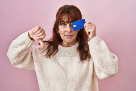 Foto de Mujer hispana de mediana edad sosteniendo el ojo de la tarjeta de crédito con la cara enojada, signo negativo que muestra aversión con los pulgares hacia abajo, concepto de rechazo - Imagen libre de derechos