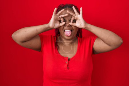 Photo for African american woman with braided hair standing over red background doing ok gesture like binoculars sticking tongue out, eyes looking through fingers. crazy expression. - Royalty Free Image
