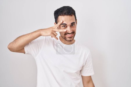 Téléchargez les photos : Bel homme hispanique debout sur fond blanc faisant symbole de paix avec les doigts sur le visage, souriant joyeux montrant la victoire - en image libre de droit