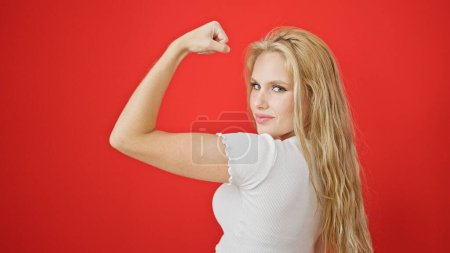 Foto de Joven mujer rubia sonriendo confiada haciendo un gesto fuerte con el brazo sobre el fondo rojo aislado - Imagen libre de derechos