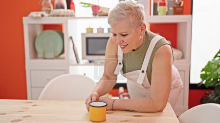 Téléchargez les photos : Femme aux cheveux gris du moyen âge buvant du café appuyé sur la table dans la salle à manger - en image libre de droit