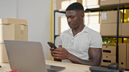 Téléchargez les photos : Homme afro-américain ecommerce travailleur d'affaires en utilisant un smartphone et un ordinateur portable au bureau - en image libre de droit