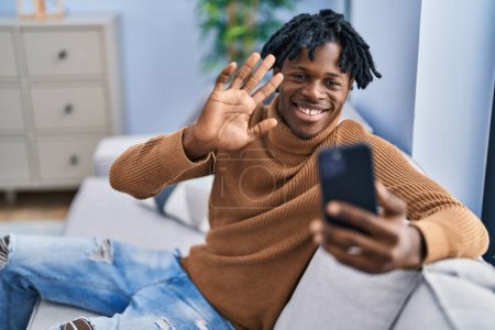 Foto de Joven hombre africano con rastas usando smartphone haciendo videollamada luciendo positivo y feliz de pie y sonriendo con una sonrisa confiada mostrando los dientes - Imagen libre de derechos