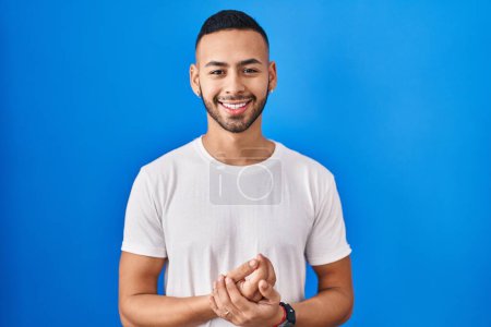 Téléchargez les photos : Jeune homme hispanique debout sur fond bleu avec les mains jointes et les doigts croisés souriant détendu et joyeux. succès et optimisme - en image libre de droit