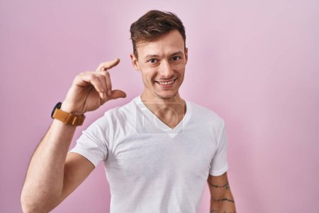 Téléchargez les photos : Homme caucasien debout sur fond rose geste souriant et confiant avec la main faisant signe de petite taille avec les doigts regardant et la caméra. concept de mesure. - en image libre de droit