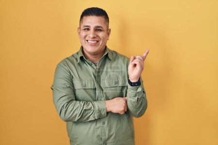 Photo for Hispanic young man standing over yellow background with a big smile on face, pointing with hand finger to the side looking at the camera. - Royalty Free Image