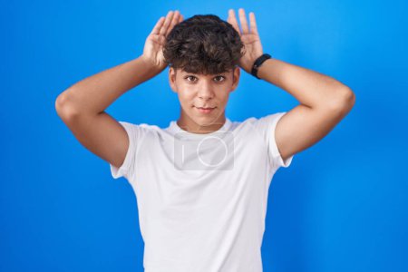 Photo for Hispanic teenager standing over blue background doing bunny ears gesture with hands palms looking cynical and skeptical. easter rabbit concept. - Royalty Free Image