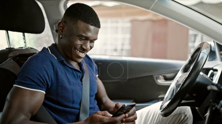 Téléchargez les photos : Afro-Américain utilisant smartphone assis sur la voiture à la rue - en image libre de droit