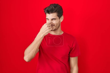 Téléchargez les photos : Jeune homme hispanique debout sur fond rouge sentant quelque chose de puant et dégoûtant, odeur intolérable, retenant souffle avec les doigts sur le nez. mauvaise odeur - en image libre de droit