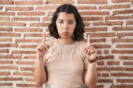 Photo for Young hispanic woman standing over bricks wall pointing up looking sad and upset, indicating direction with fingers, unhappy and depressed. - Royalty Free Image