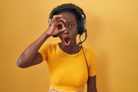 Photo for African woman with curly hair standing over yellow background wearing headphones doing ok gesture shocked with surprised face, eye looking through fingers. unbelieving expression. - Royalty Free Image