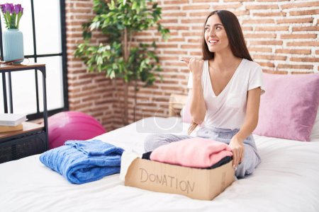 Photo for Young hispanic woman putting used clothes into donations box sitting on the bed smiling with happy face looking and pointing to the side with thumb up. - Royalty Free Image