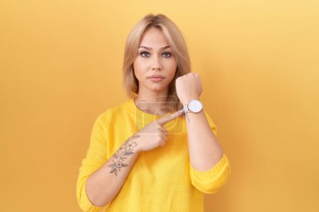 Téléchargez les photos : Jeune femme caucasienne portant un pull jaune pressé pointant pour regarder le temps, l'impatience, regardant la caméra avec une expression détendue - en image libre de droit
