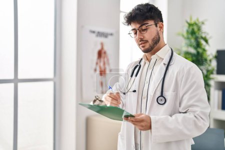 Téléchargez les photos : Jeune homme hispanique portant un uniforme de médecin rédigeant un rapport médical à la clinique - en image libre de droit
