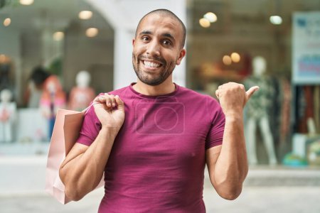Photo for Hispanic man with beard holding shopping bags outdoors pointing thumb up to the side smiling happy with open mouth - Royalty Free Image