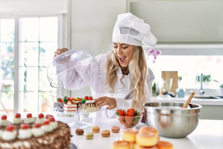 Téléchargez les photos : Jeune femme portant cuisinière plateau d'ouverture uniforme avec des gâteaux à la cuisine - en image libre de droit