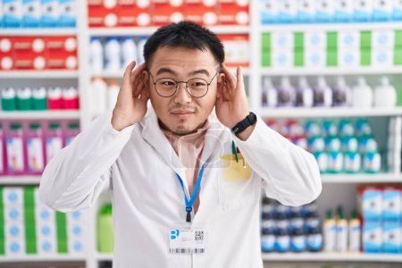 Foto de Un joven chino que trabaja en una farmacia tratando de escuchar ambos gestos, curioso por los chismes. problema auditivo, sordo - Imagen libre de derechos