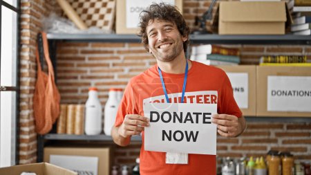 Téléchargez les photos : Jeune homme hispanique bénévole souriant confiant tenant faire un don maintenant bannière au centre de charité - en image libre de droit