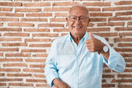 Téléchargez les photos : Homme âgé avec les cheveux gris debout sur le mur de briques faisant des pouces heureux geste avec la main. approuvant l'expression regardant la caméra montrant le succès. - en image libre de droit