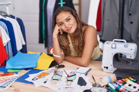 Téléchargez les photos : Jeune belle femme hispanique tailleur souriant confiant assis sur la table au magasin de tailleur - en image libre de droit