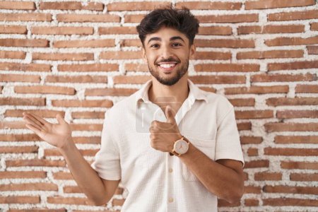 Foto de Hombre árabe con barba de pie sobre ladrillos fondo de la pared mostrando la palma de la mano y haciendo buen gesto con los pulgares hacia arriba, sonriendo feliz y alegre - Imagen libre de derechos