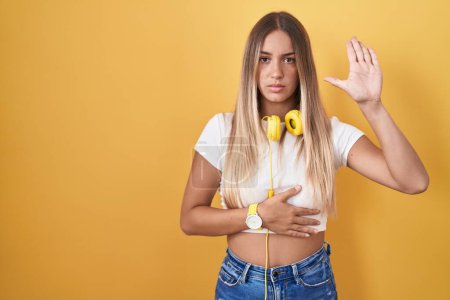 Photo for Young blonde woman standing over yellow background wearing headphones swearing with hand on chest and open palm, making a loyalty promise oath - Royalty Free Image
