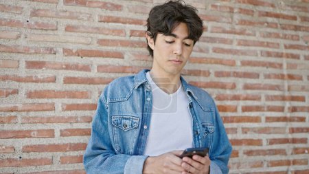 Téléchargez les photos : Jeune homme hispanique utilisant un smartphone avec une expression sérieuse sur fond de briques isolées - en image libre de droit