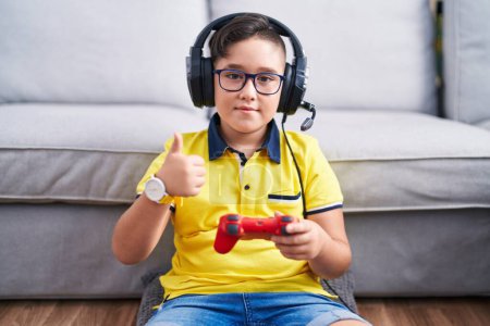 Téléchargez les photos : Jeune enfant hispanique jouant contrôleur de tenue de jeu vidéo portant des écouteurs faisant des pouces heureux geste avec la main. approuvant l'expression regardant la caméra montrant le succès. - en image libre de droit