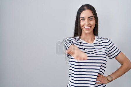Foto de Joven morena vistiendo camiseta a rayas sonriente alegre ofreciendo la palma de la mano dando asistencia y aceptación. - Imagen libre de derechos