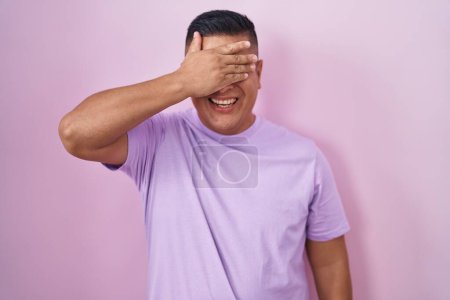 Téléchargez les photos : Jeune homme hispanique debout sur fond rose souriant et riant avec la main sur le visage couvrant les yeux pour la surprise. concept aveugle. - en image libre de droit