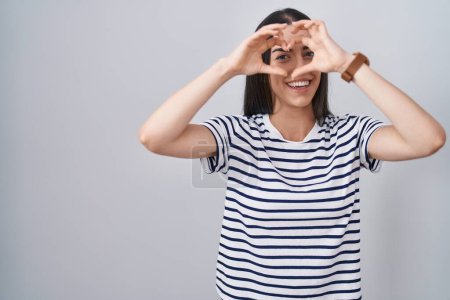 Téléchargez les photos : Jeune femme brune portant un t-shirt rayé en forme de coeur avec la main et les doigts souriant en regardant à travers le signe - en image libre de droit