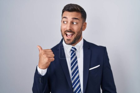 Foto de Hombre hispano guapo vistiendo traje y corbata sonriendo con la cara feliz mirando y señalando a un lado con el pulgar hacia arriba. - Imagen libre de derechos
