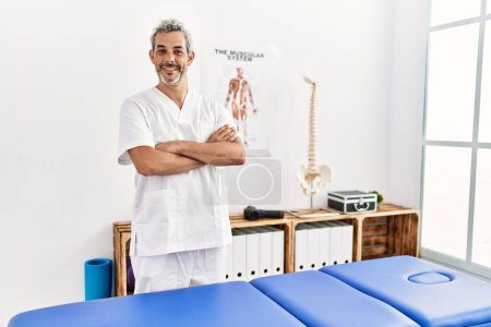 Foto de Pisioterapeuta hombre de pelo gris de mediana edad sonriendo confiado de pie con los brazos cruzados gesto en la clínica de rehabilitación - Imagen libre de derechos