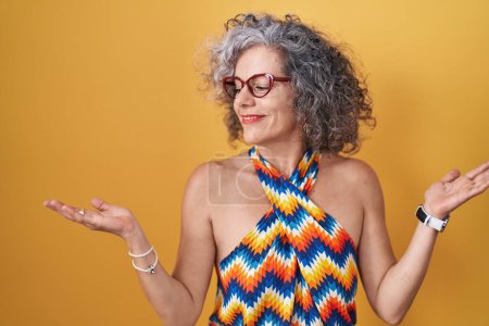 Photo for Middle age woman with grey hair standing over yellow background smiling showing both hands open palms, presenting and advertising comparison and balance - Royalty Free Image