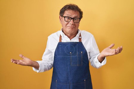 Photo for Middle age hispanic man wearing professional cook apron clueless and confused expression with arms and hands raised. doubt concept. - Royalty Free Image