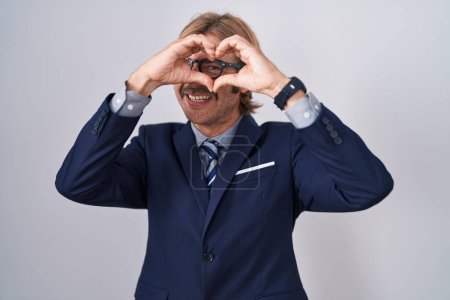 Foto de Hombre caucásico con bigote usando ropa de negocios haciendo forma de corazón con la mano y los dedos sonriendo mirando a través de signo - Imagen libre de derechos