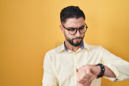 Téléchargez les photos : Jeune homme hispanique portant des vêtements de travail et des lunettes vérifiant l'heure sur la montre-bracelet, détendu et confiant - en image libre de droit