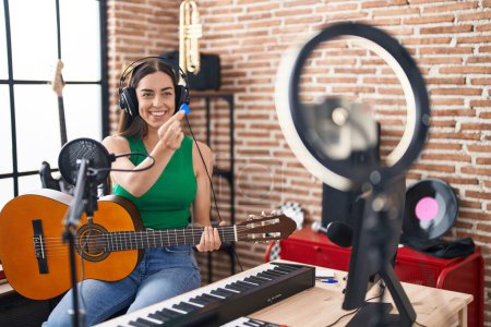 Foto de Young hispanic woman musician recording classical guitar lesson by the smartphone at music studio - Imagen libre de derechos