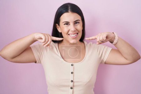 Foto de Mujer hispana joven de pie sobre fondo rosa sonriendo alegre mostrando y señalando con los dedos dientes y la boca. concepto de salud dental. - Imagen libre de derechos