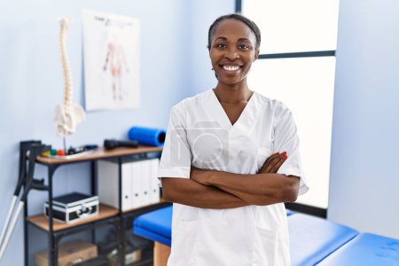 Téléchargez les photos : Femme afro-américaine pysiothérapeute souriant confiant debout avec les bras croisés geste à la clinique de réadaptation - en image libre de droit