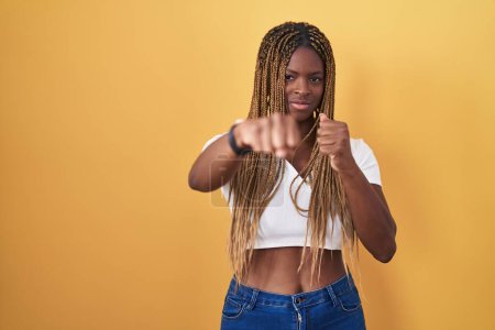 Photo for African american woman with braided hair standing over yellow background punching fist to fight, aggressive and angry attack, threat and violence - Royalty Free Image