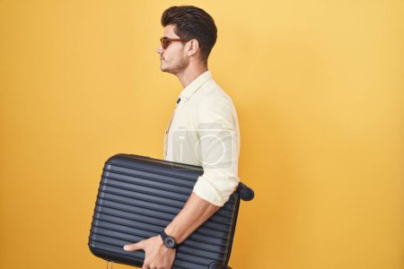 Foto de Joven hombre hispano sosteniendo maleta va de vacaciones de verano buscando a un lado, relajarse pose perfil con cara natural y sonrisa confiada. - Imagen libre de derechos