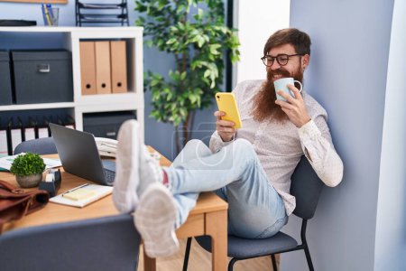 Téléchargez les photos : Jeune rouquine homme d'affaires en utilisant smartphone boire du café au bureau - en image libre de droit