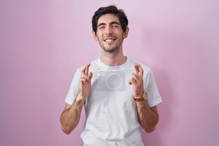 Téléchargez les photos : Jeune homme hispanique debout sur fond rose geste doigt croisé souriant avec espoir et les yeux fermés. chance et concept superstitieux. - en image libre de droit