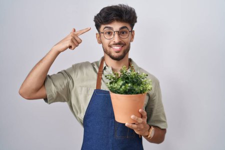 Téléchargez les photos : Homme arabe avec barbe tenant pot de plante verte souriant pointant vers la tête avec un doigt, grande idée ou pensée, bonne mémoire - en image libre de droit