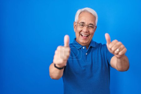 Foto de Hombre de mediana edad con el pelo gris de pie sobre fondo azul aprobando hacer un gesto positivo con la mano, pulgares hacia arriba sonriendo y feliz por el éxito. gesto ganador. - Imagen libre de derechos